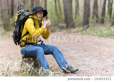 一名在森林中徒步旅行的男子坐在樹樁上休息。 一名在森林中徒步旅行的男子坐在樹樁上休息。 143051411