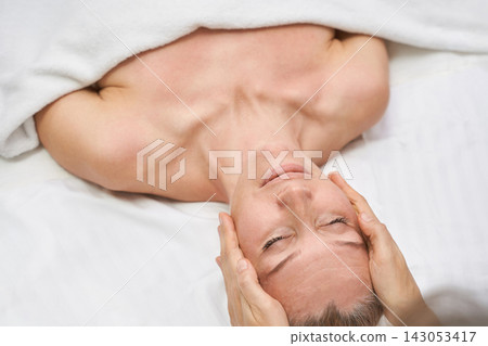 Woman Receiving Facial Massage With Eyes Closed Under White Blanket In Calm Spa Treatment Room Woman Receiving Facial Massage With Eyes Closed Under White Blanket In Calm Spa Treatment Room 143053417
