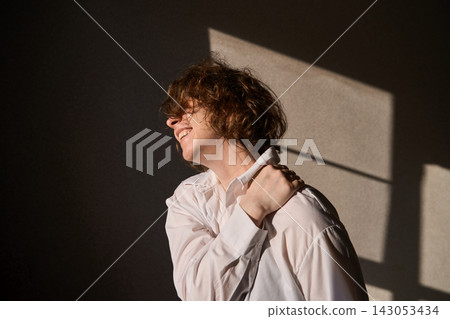 Young Woman Clutching Shoulder In Sunlit Interior With Sharp Window Shadows Across Dark Wall Young Woman Clutching Shoulder In Sunlit Interior With Sharp Window Shadows Across Dark Wall 143053434