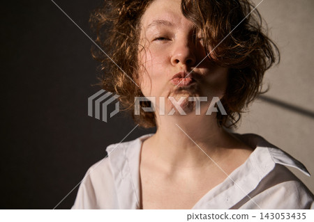 Woman Puckering Lips With Closed Eyes In White Shirt Under Warm Side Light In Studio Portrait Woman Puckering Lips With Closed Eyes In White Shirt Under Warm Side Light In Studio Portrait 143053435