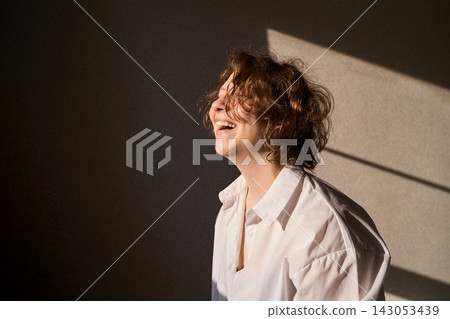 Smiling Curly Haired Woman In White Shirt Posing In Warm Sunlight Against Beige Wall With Sharp Smiling Curly Haired Woman In White Shirt Posing In Warm Sunlight Against Beige Wall With Sharp 143053439