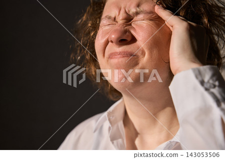 Adult Woman Pressing Temple And Squeezing Eyes Shut In White Shirt Under Dramatic Side Light Adult Woman Pressing Temple And Squeezing Eyes Shut In White Shirt Under Dramatic Side Light 143053506
