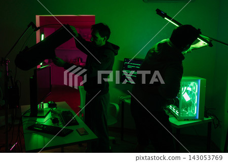 Two technicians prepare for filming or streaming in dark room. Men adjust professional lights near computers with code on screens. Green neon glow creates cinematic, mysterious hacker atmosphere. Two technicians prepare for filming or streaming in dark room. Men adjust professional lights near computers with code on screens. Green neon glow creates cinematic, mysterious hacker atmosphere. 143053769