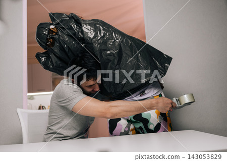 Man playfully wraps friend in large black trash bag with packaging tape in quirky indoor prank, generating laughter and silly, surprising bonding moment. Man playfully wraps friend in large black trash bag with packaging tape in quirky indoor prank, generating laughter and silly, surprising bonding moment. 143053829