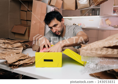 Man with dark hair preparing small yellow cardboard box with protective padding for delivery. Surrounded by various packaging materials like crumpled paper and bubble wrap. Focusing on sending items. Man with dark hair preparing small yellow cardboard box with protective padding for delivery. Surrounded by various packaging materials like crumpled paper and bubble wrap. Focusing on sending items. 143053831