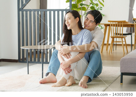 一位年輕的日本男子從背後擁抱了一位坐在客廳裡的年輕日本女子。 一位年輕的日本男子從背後擁抱了一位坐在客廳裡的年輕日本女子。 143053950