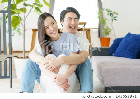 一位年輕的日本男子從背後擁抱了一位坐在客廳裡的年輕日本女子。 一位年輕的日本男子從背後擁抱了一位坐在客廳裡的年輕日本女子。 143053951