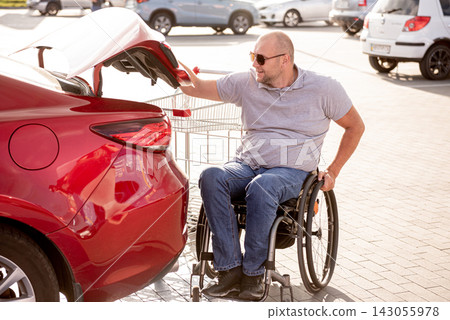 Adult disabled man in a wheelchair puts purchases in the trunk of a car in a supermarket parking lot 143055978