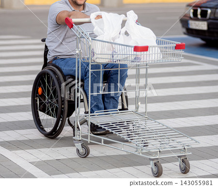 Disabled man in wheelchair pushing cart in front of himself at supermarket parking Disabled man in wheelchair pushing cart in front of himself at supermarket parking 143055981