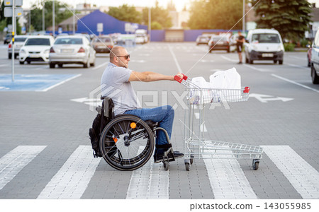 Disabled man in wheelchair pushing cart in front of himself at supermarket parking Disabled man in wheelchair pushing cart in front of himself at supermarket parking 143055985
