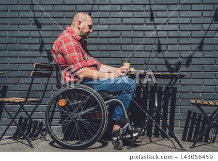 Freelancer with a physical disability in a wheelchair working at the street cafe Freelancer with a physical disability in a wheelchair working at the street cafe 143056459