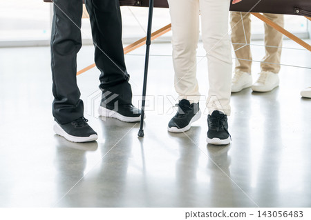 一位老年人正在接受使用拐杖的步行訓練。護理人員為其提供復健和防跌倒方面的支援。 一位老年人正在接受使用拐杖的步行訓練。護理人員為其提供復健和防跌倒方面的支援。 143056483