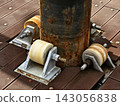 Rusty metal pipe support with three roller wheels on weathered wooden deck in sunlight 143056838