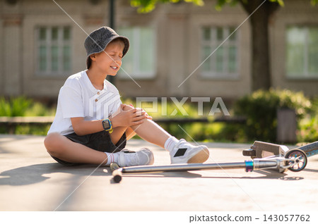 Boy in a hat sitting on the ground near the scooter after a fall down Boy in a hat sitting on the ground near the scooter after a fall down 143057762