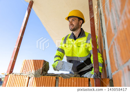 Happy builder wearing yellow hard hat holding trowel checking alignment Happy builder wearing yellow hard hat holding trowel checking alignment 143058417