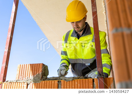 Male mason wearing helmet applying cement mixture with trowel Male mason wearing helmet applying cement mixture with trowel 143058427
