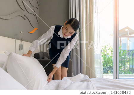 Female hotel housekeeper in navy uniform making bed with fresh white linens and pillows in modern hotel guest room. Smiling Asian woman demonstrating quality hospitality service Female hotel housekeeper in navy uniform making bed with fresh white linens and pillows in modern hotel guest room. Smiling Asian woman demonstrating quality hospitality service 143058483