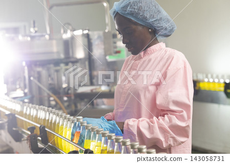 Black woman inspecting drink quality with rows of glass bottles on the automated conveyor system in manufacturing assembly line. Black woman inspecting drink quality with rows of glass bottles on the automated conveyor system in manufacturing assembly line. 143058731
