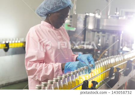 Black woman inspecting drink quality with rows of glass bottles on the automated conveyor system in manufacturing assembly line. 143058732