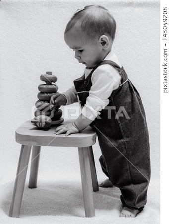 Black and white art portrait of cute baby standing near small wooden stool and playing with pyramid toy. Vintage childhood style, first standing steps, and nostalgic family moments Black and white art portrait of cute baby standing near small wooden stool and playing with pyramid toy. Vintage childhood style, first standing steps, and nostalgic family moments 143059208