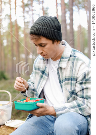 Man eating a healthy lunch from a plastic container in the forest, enjoying an outdoor meal during a hiking trip, sitting on a log with a fork and snacks. Man eating a healthy lunch from a plastic container in the forest, enjoying an outdoor meal during a hiking trip, sitting on a log with a fork and snacks. 143062966