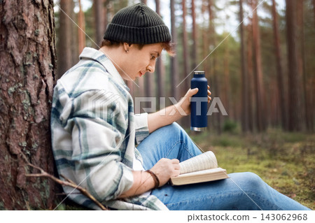 Man reading a book and drinking from a thermos in the forest, enjoying digital detox, mindfulness, nature therapy, stress relief, mental wellbeing, outdoor leisure, and relaxation. Man reading a book and drinking from a thermos in the forest, enjoying digital detox, mindfulness, nature therapy, stress relief, mental wellbeing, outdoor leisure, and relaxation. 143062968