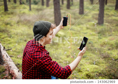 Man charging a smartphone with a portable powerbank in the forest, staying connected outdoors during hiking and travel. Man charging a smartphone with a portable powerbank in the forest, staying connected outdoors during hiking and travel. 143062969