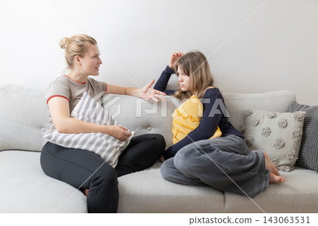 Two young women engaged in a conversation on a light gray sofa, one gesturing with a pillow while the other listens attentively, casual home interior setting Two young women engaged in a conversation on a light gray sofa, one gesturing with a pillow while the other listens attentively, casual home interior setting 143063531