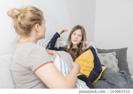 Two young women engaged in conversation on a couch, one holding a pillow, with decorative cushions and a light-colored wall in a cozy indoor setting Two young women engaged in conversation on a couch, one holding a pillow, with decorative cushions and a light-colored wall in a cozy indoor setting 143063533