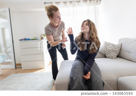 Caucasian woman talking to teenage girl sitting on a gray sofa in a bright living room, with a light-colored rug and furniture visible in the background Caucasian woman talking to teenage girl sitting on a gray sofa in a bright living room, with a light-colored rug and furniture visible in the background 143063593