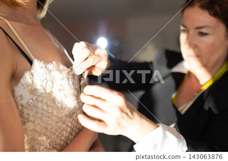 Female fashion designer adjusts intricate wedding dress on model, showcasing detailed beadwork and lace in a well-lit fitting room environment Female fashion designer adjusts intricate wedding dress on model, showcasing detailed beadwork and lace in a well-lit fitting room environment 143063876