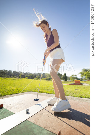 Girl plays mini golf in sunny park on a clear day while focusing on hitting the ball towards the hole Girl plays mini golf in sunny park on a clear day while focusing on hitting the ball towards the hole 143064287