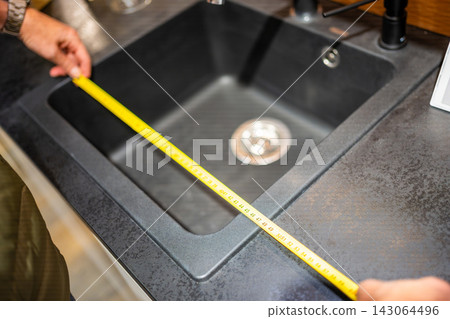 Man measures kitchen sink with tape measure in furniture showroom. Kitchen renovation and installation planning with selective focus and soft background Man measures kitchen sink with tape measure in furniture showroom. Kitchen renovation and installation planning with selective focus and soft background 143064496