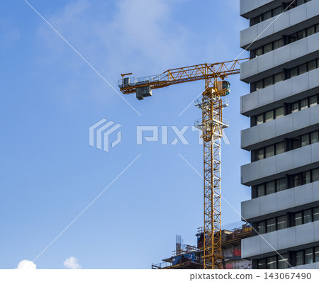 Low angle view of a yellow tower crane operating next to a modern high-rise building under a blue sky. Low angle view of a yellow tower crane operating next to a modern high-rise building under a blue sky. 143067490