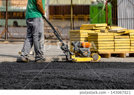 A man operates an asphalt compactor on a road construction site. Nearby, stacks of paving stones stand ready for installation A man operates an asphalt compactor on a road construction site. Nearby, stacks of paving stones stand ready for installation 143067821