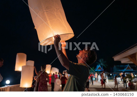 Young man releasing glowing paper sky lantern at night during festival, warm flame inside lantern lighting his hands, night sky background with soft bokeh lights and celebratory atmosphere Young man releasing glowing paper sky lantern at night during festival, warm flame inside lantern lighting his hands, night sky background with soft bokeh lights and celebratory atmosphere 143068150