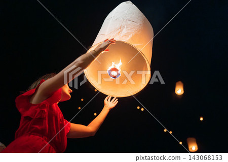 Woman in red dress releasing illuminated paper lantern into night sky during cultural festival, glowing lantern launch with warm orange light, upward gaze and distant floating lanterns 143068153