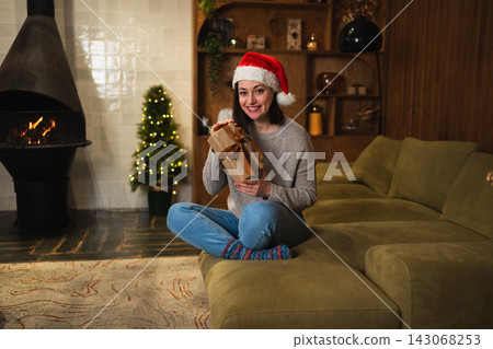 Merry Christmas. Young woman in Santa hat unpacking open up gift box near Christmas tree. Girl in living room with Christmas tree, fireplace opening gift box with surprise face. Christmas eve at home Merry Christmas. Young woman in Santa hat unpacking open up gift box near Christmas tree. Girl in living room with Christmas tree, fireplace opening gift box with surprise face. Christmas eve at home 143068253