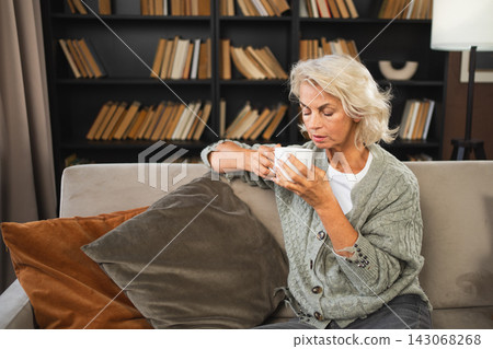 Good morning autumn winter fall vibe. Comfort relaxed senior old elderly woman drinking hot beverage tea coffee in sofa at home. Mature woman resting enjoy calm morning at home alone. Pension concept 143068268