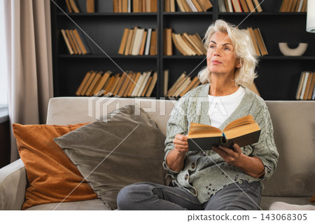 Comfort relaxed senior old elderly woman reading book at home. Mature woman resting enjoy calm day sitting on sofa at home alone. Adult lady spending free time keeping book smiling. Pension concept Comfort relaxed senior old elderly woman reading book at home. Mature woman resting enjoy calm day sitting on sofa at home alone. Adult lady spending free time keeping book smiling. Pension concept 143068305