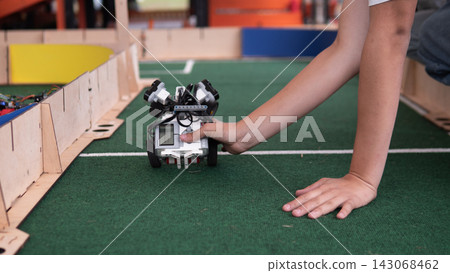 Child hand steadies compact wheeled robot on green indoor track beside wooden barriers, showing hands on robotics learning, coding practice, STEM education, and teamwork in classroom challenge area Child hand steadies compact wheeled robot on green indoor track beside wooden barriers, showing hands on robotics learning, coding practice, STEM education, and teamwork in classroom challenge area 143068462
