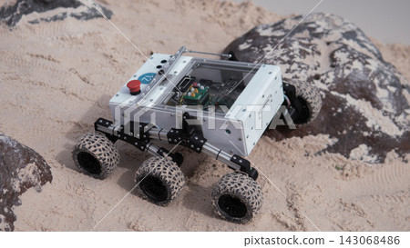 Compact four wheeled exploration robot with white protective body, clear top panel, exposed electronics and rugged suspension crawling over sandy terrain beside dark rocks during off road mobility Compact four wheeled exploration robot with white protective body, clear top panel, exposed electronics and rugged suspension crawling over sandy terrain beside dark rocks during off road mobility 143068486
