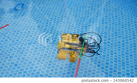 Yellow robotic pool device with black cables floating in clear blue water above hexagonal tiles, suggesting automated underwater cleaning, inspection, and maintenance testing for pool hygiene Yellow robotic pool device with black cables floating in clear blue water above hexagonal tiles, suggesting automated underwater cleaning, inspection, and maintenance testing for pool hygiene 143068542
