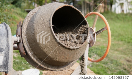 Rotating cement mixer drum packed with wet concrete and gravel, captured outdoors during construction work. Rusted metal surface, rough texture, shallow depth, green site background Rotating cement mixer drum packed with wet concrete and gravel, captured outdoors during construction work. Rusted metal surface, rough texture, shallow depth, green site background 143068567