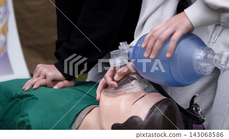 Close view shows hands pressing training mannequin chest while another person holds ventilation mask and bag valve device for emergency resuscitation practice in outdoor setting, highlighting CPR Close view shows hands pressing training mannequin chest while another person holds ventilation mask and bag valve device for emergency resuscitation practice in outdoor setting, highlighting CPR 143068586