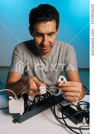 Vertical portrait of man holding multiple power plugs plugging cables and adapters into overcrowded power strip on desk, creating tangled, messy cluster of wires and chargers. Vertical portrait of man holding multiple power plugs plugging cables and adapters into overcrowded power strip on desk, creating tangled, messy cluster of wires and chargers. 143068828