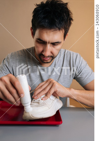Vertical portrait of focused young man applying white protective paint to dirty sneaker, providing shoe care, renewal and restoration, cleaning footwear and demonstrating maintenance process. 143068910