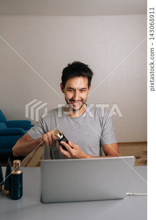 Vertical portrait of smiling man vlogging about beauty and skincare, doing online review of cosmetic products using laptop, engaging with audience through streaming for social media broadcast. 143068911