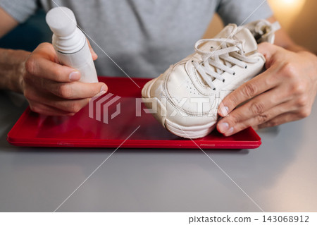 Cropped shot of unrecognizable man holding using bottle of shoe whitener and painting scuffed surface of dirty white sneaker, illustrating process of cleaning, recoloring, and restoring footwear. Cropped shot of unrecognizable man holding using bottle of shoe whitener and painting scuffed surface of dirty white sneaker, illustrating process of cleaning, recoloring, and restoring footwear. 143068912