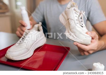 Close-up cropped shot of unrecognizable man holding shoe cleaner and white sneaker, preparing for cleaning and maintenance, focusing on domestic tasks and product use for footwear restoration. Close-up cropped shot of unrecognizable man holding shoe cleaner and white sneaker, preparing for cleaning and maintenance, focusing on domestic tasks and product use for footwear restoration. 143068915